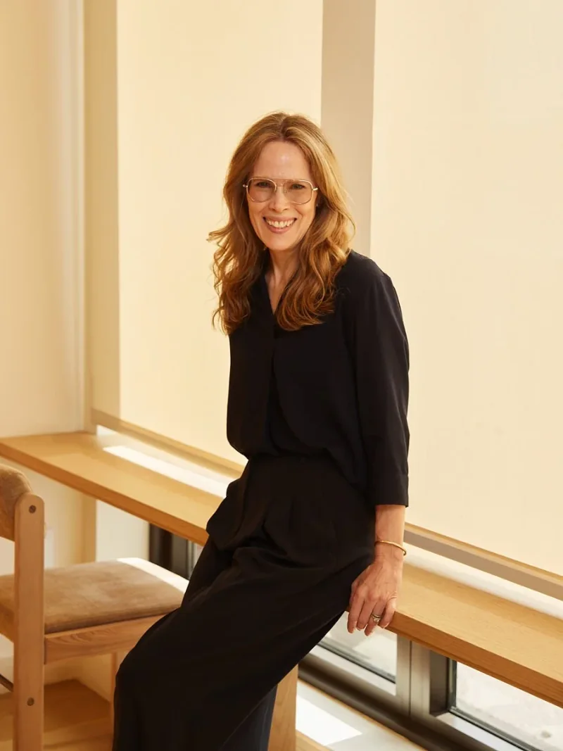Woman in a black outfit and glasses seated beside a sunlit window.
