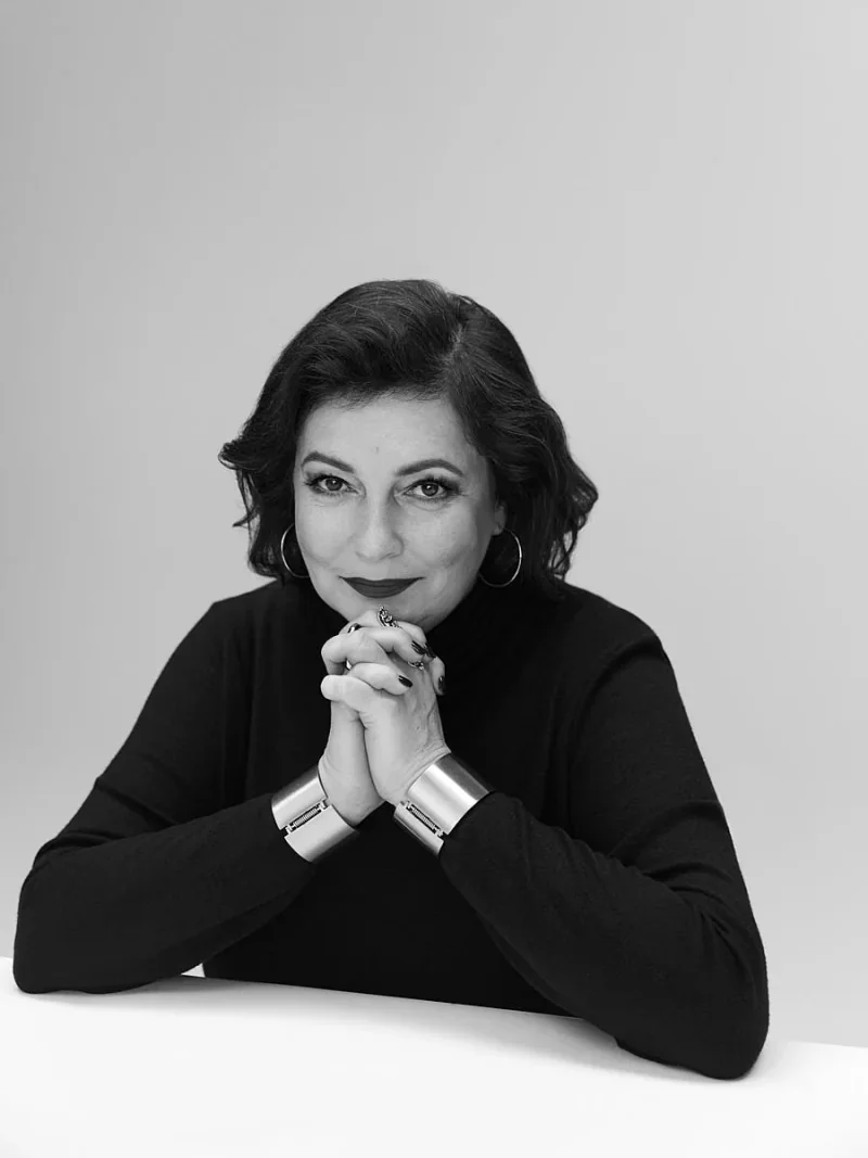 Black and white portrait of a woman in a dark top seated at a table with her hands beneath her chin.
