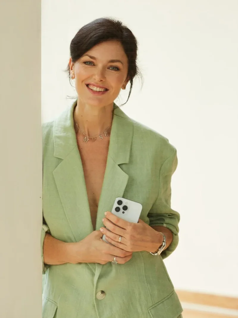 Woman in a light green blazer holding a phone beside a window.