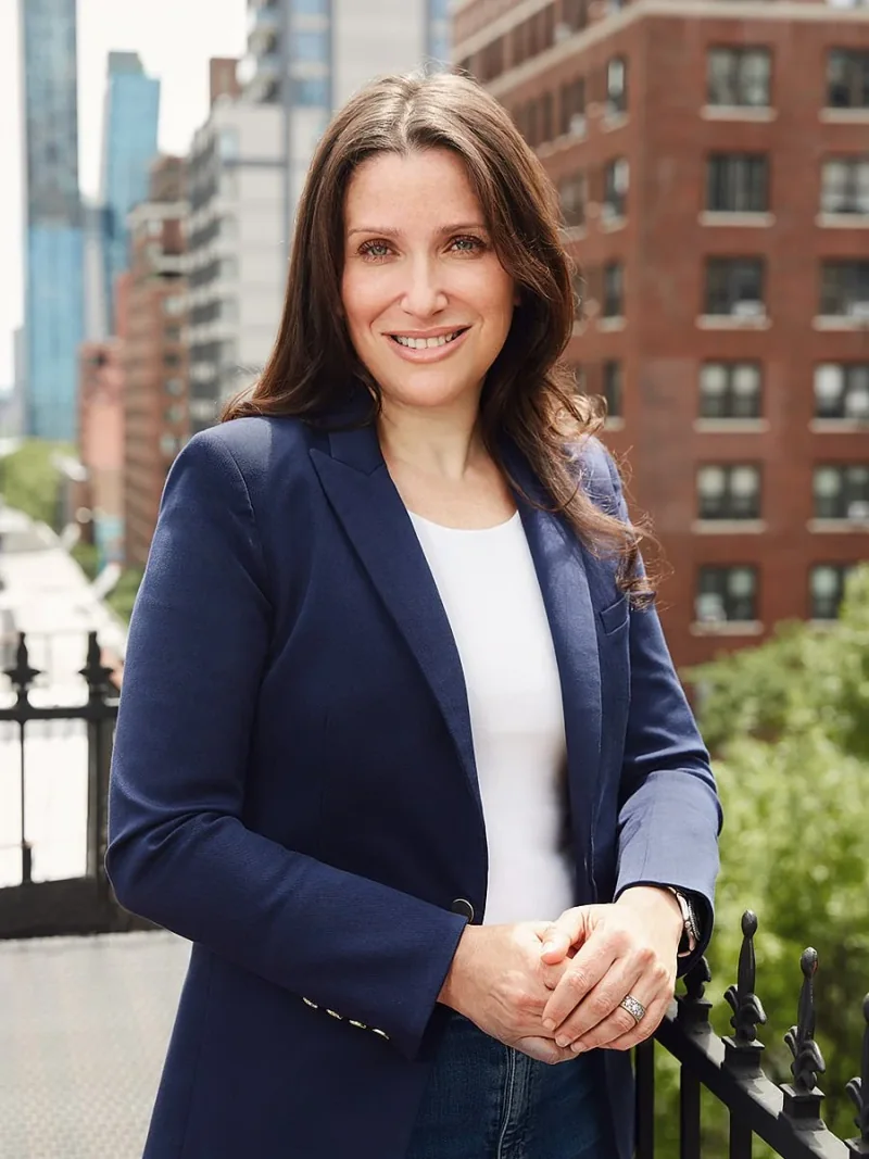 Woman in a navy blazer standing on a balcony with city buildings behind her.