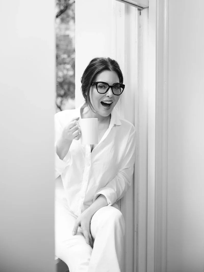 Black and white portrait of a woman in glasses and white clothing seated by a window holding a mug.